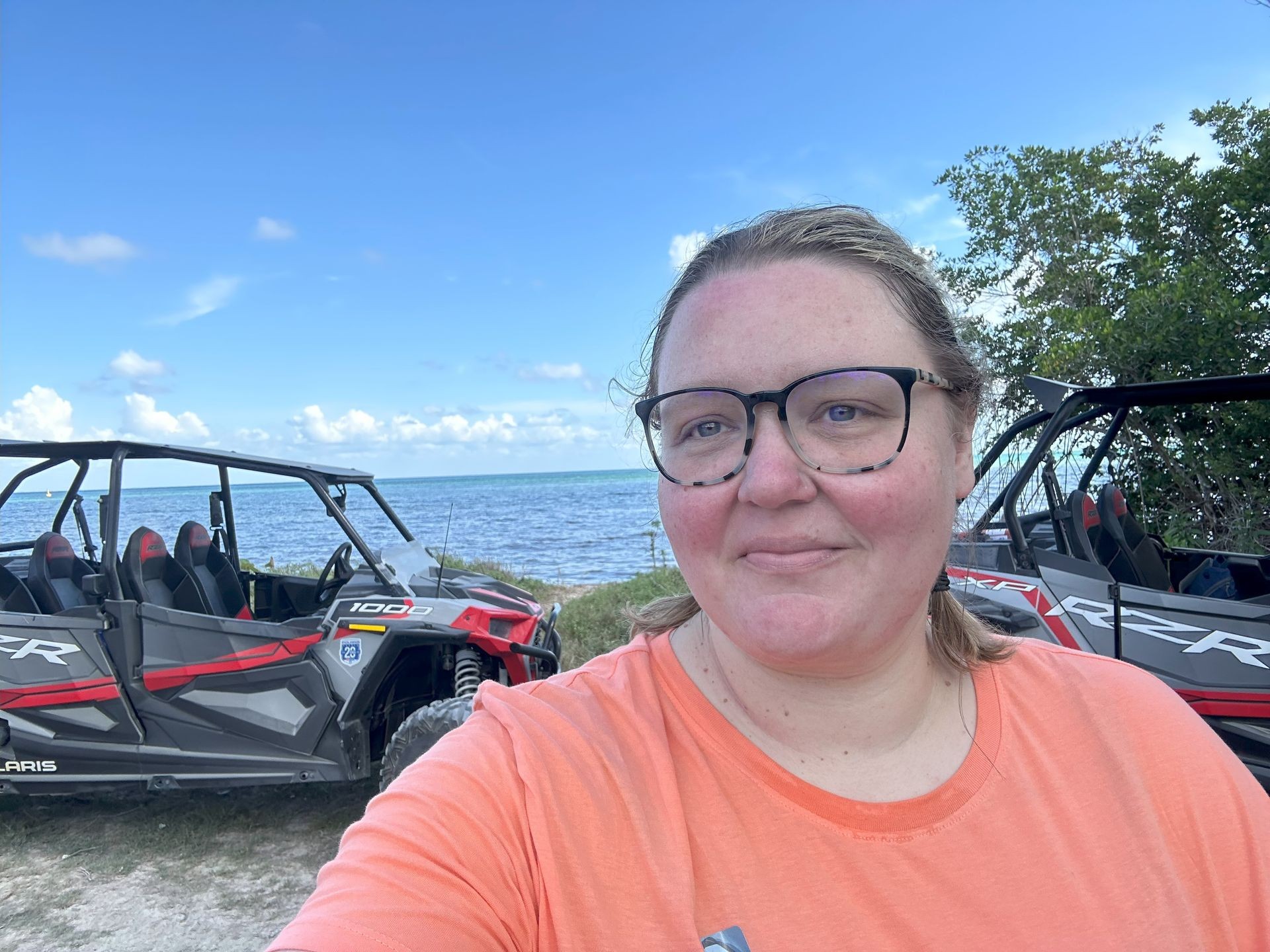 Person in glasses with side-by-side off-road vehicles near a scenic ocean view.